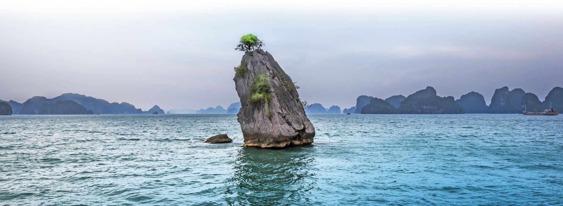 Ein Felsen steht mittig ein einem großen See - drumherum sind kleiner Berge und ein Schiff